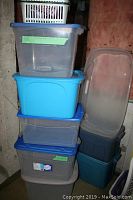 Stack of various plastic storage totes and small baskets, showing different sizes and colors, mostly clear and blue with lids. Includes under-bed bin without lid leaning on stack.