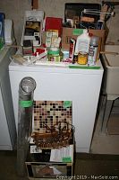 Overview of many items on and in front of a white appliance, showing pipe, tile, light fixture part, paint cans, and assorted smaller items on top and shelves.