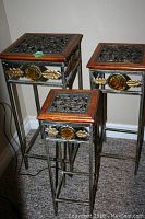 Three nesting plant stands with ornate metal scrollwork tops, wood borders, and decorative amber glass medallions with gold leaf details on sides.