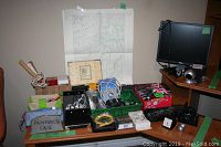 Wide view of table with assorted electronics, office supplies, craft supplies, and map or blueprint pinned behind.