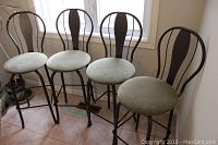 Four matching swivel bar stools with upholstered green seats and black metal frames, arranged in a row near a window on a tiled floor.