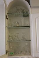 Full view of the glass cabinet with four glass shelves holding assorted glassware including decanters, vases, candle holders, and bowls.