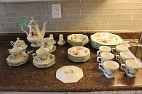 Image showing full dish set on counter including teapot, sugar bowl with lid and broken handle, creamer, salt and pepper shakers, cups, saucers, salad and dinner plates. White porcelain with colorful fruit design.