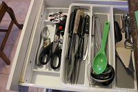 Kitchen drawer containing assorted knives, scissors, metal corkscrew, green plastic spoon, and metal measuring spoons organized in white plastic drawer inserts.