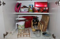 Kitchen cabinet interior showing various kitchen items including knife block with knives, cutting boards, lunch bags, cookie cutters, jars, napkin holder, and other kitchen accessories