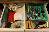 Overview of drawer with cutlery tray containing forks, knives, spoons, metal BBQ skewers with wooden handles, red Starfrit kitchen scale, green marble rolling pin, and Pampered Chef mandolin slicer.