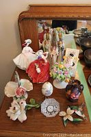 Photo showing the full assortment of porcelain figurines, clock, jug, and music box on a wooden surface with mirror reflection behind.
