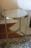 Round glass top end table with metal frame, showing table supported on four legs connected by a curved metal brace, displayed against a white brick wall and hardwood floor with patterned rug.