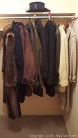 Photo of various outerwear items hanging on rack including full fur coat, leather coats, and trench coat with hat on top shelf