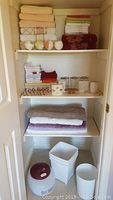 Closet shelves with folded assorted towels, face cloths, decorative rose shower curtain hooks, bath bombs, ceramic bathroom accessory sets, bath mats, two trash cans, and Sunbeam vaporizer.