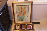 Photo showing one large framed floral watercolor and three smaller framed floral foil artworks arranged on the floor.