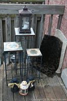 Photo showing three metal plant stands with mosaic tile tops in three sizes, four lanterns of varying sizes and designs, and two rolled mats leaning against the wall.