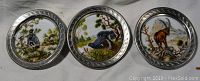 Three round china coasters with painted scenes of birds and deer each in an ornate embossed metal rim.