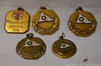 Photo showing five gold-tone rowing regatta medallions, front view. Includes one octagonal 1912 Champion Regatta medallion and four round medallions with flag and laurel wreath.