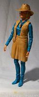 Full body side view of Jane West action figure wearing blue shirt, tan vest and skirt, and tan hat