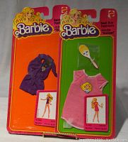 Photo showing two Barbie Best Buy Fashions outfits in sealed blister cards, one purple dress and one pink dress with a mirror accessory.