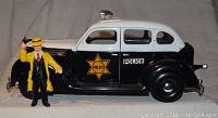 Side view of black and white Dick Tracy police car toy with Dick Tracy figure standing beside it. Car has police star decals and black wheels.