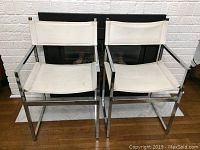 Pair of vintage chairs with chrome frames and white seats and backs placed side by side against a fireplace.