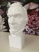 White ceramic bust of Abraham Lincoln with detailed facial features and beard, on a square pedestal base, photographed against floral background.