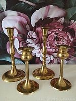Four polished brass candle holders arranged in front of a floral backdrop, showing full height and shape differences