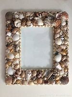 Top-down photo showing the full square shell photo frame with a clear center for photo display. The frame is densely decorated with various shells and small beads.
