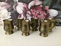 Photo showing six brass mugs arranged in a group in front of floral background, highlighting hammered texture and vintage style.