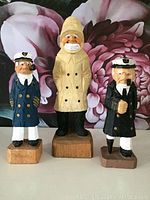 Front view of three wood carved figures on wooden bases, against floral background.