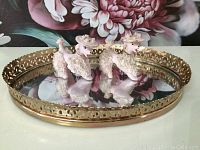 Two white vintage poodle figurines with textured fur and floral hats sitting on an oval mirror tray with gold trim, reflecting below.