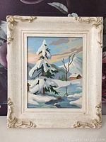 Full view of vintage paint by number painting in white ornate frame showing a winter scene with snow, pine tree, creek, and cabin.