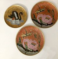 Three decorative brass plates: two with pink flowers and dragonflies on orange background, one with black swan on water on gold background.
