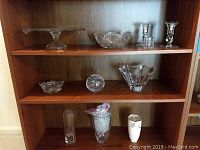 Wooden shelf with multiple clear crystal glassware items including cake plate, cut glass bowl, two crystal candlesticks, glass votive holder, and decorative vases.