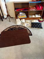 Wide view showing shelving unit with folded table linens in various colors, stacks of doilies and napkins, vernacular pillow with fleur pattern, brown kitchen mat, and semicircle patterned door mat on floor.