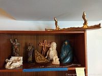 Full view of shelf displaying multiple religious figurines and wall shelves including one blue-robed Madonna figurine, ivory Mary and child, and several wooden shelves above