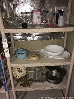 Full view of shelf showing various glass and ceramic serving dishes, metal trivets, and cookware items arranged on three shelves.