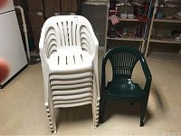 Stacked group of eight white plastic chairs next to two green plastic chairs on basement floor.