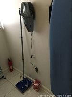 Photo showing the Dirt Devil vacuum mounted on a wall charger, blue floor sweeper with a long handle, and foldable ironing board nearby