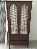 Front view of a wooden armoire with two closed door panels featuring lattice design and two bottom drawers with brass handles.
