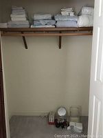 Closet shelf with stacked linens and items on closet floor including vanity mirror, hairdryer, and faucet