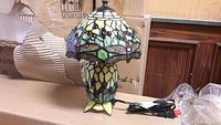 Loose Tiffany-style stained glass table lamp showing colorful multi-paned glass shade and matching base with power cord and inline switch.