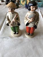 Two vintage Asian figurines: a boy with green pants and conical hat playing an instrument, and a girl with red pants holding a fan, both painted and standing on circular bases.