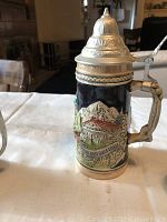 Front and side view of ceramic beer stein with relief scenic design and pewter lid in a living room setting.