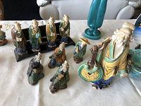 Photo of nine vintage painted mud man figurines on a white cloth, including a larger figure holding a fan and tray with coins, and eight smaller seated and standing figures with traditional attire and poses.