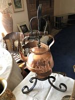 Photo shows a round copper tea kettle with attached handles sitting on a decorative wrought iron stand with curled legs and a copper Sterno holder beneath the kettle for heating.