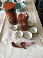 Photo showing red lacquer hexagonal sake decanter, five small ceramic sake cups with white inside and decorative exterior, and two red ornamental ceramic spoons on a white cloth
