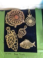 Photo of 7 assorted brass trivets with various shapes and openwork designs, displayed on black fabric with green background