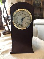 Upright wooden table clock with a rounded top, Roman numeral clock face, and brass rimmed glass cover.
