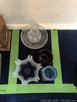 Top view of four assorted glassware items: clear bowl and decanter with etched detail, maroon glass bowl, blue frosted square vase, and ruffled blue-white art glass vase.