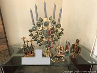 Full view of Mexican paper mache collectibles arranged on a glass table including a floral candelabra with candles, church sculpture, bird nest with birds, and angel figurine.