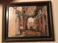 Framed Mexican street scene with arches, balconies, and three figures including one sitting and two walking on street.