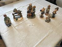 Overview photo showing all 7 German porcelain figurines by Hummel, Goebel and others on a white cloth.